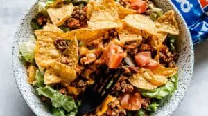 A close-up of a Dorito Taco Salad in a speckled white bowl, featuring crunchy nacho cheese Doritos, seasoned ground beef, fresh lettuce, diced tomatoes, and melted cheese, with a black fork resting on top.