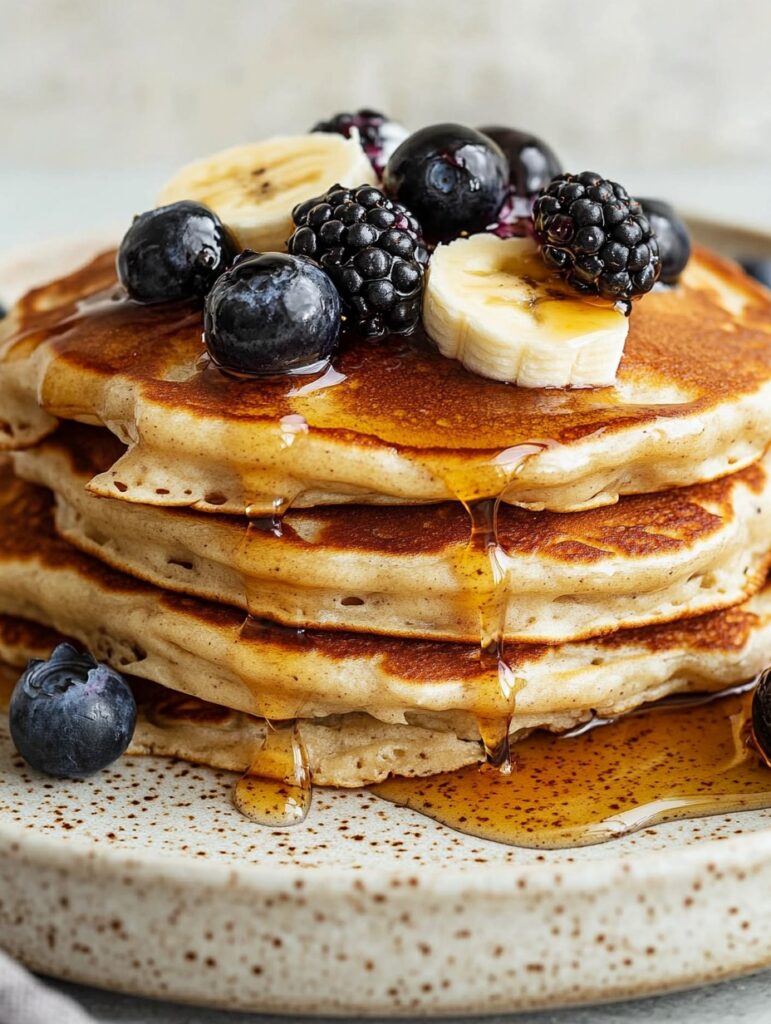 Fluffy protein pancakes topped with fresh blueberries, blackberries, banana slices, and drizzled with golden maple syrup.